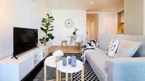 Un salon moderne avec un téléviseur à écran plat, un canapé gris, une table à manger avec des chaises et une grande plante.