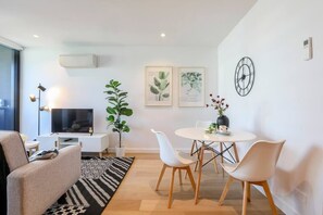 A modern living room with a flat-screen TV, a sofa, a dining table with chairs, and two framed botanical prints on the wall.
