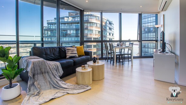 A modern living room with a large window, a sofa, a coffee table, and a dining area. - KOZYGURU | DOCKLANDS | HIGH LEVEL WATERVIEW | 1BED  |  POOL  | VDO883 (Docklands)
