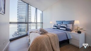 A modern bedroom with a large window, a bed with a blue and white striped duvet, a bedside table with a lamp, and a view of a cityscape. - KOZYGURU | DOCKLANDS | HIGH LEVEL WATERVIEW | 1BED  |  POOL  | VDO883 (Docklands)