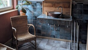 Un baño con un ventanal grande, una planta en maceta, una silla de mimbre, un armario de madera, un lavabo con espejo y paredes de azulejos.