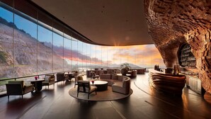 Um amplo salão com teto curvo, grandes janelas com vista para as montanhas e um espaço de bar com design exclusivo na parede de pedra.