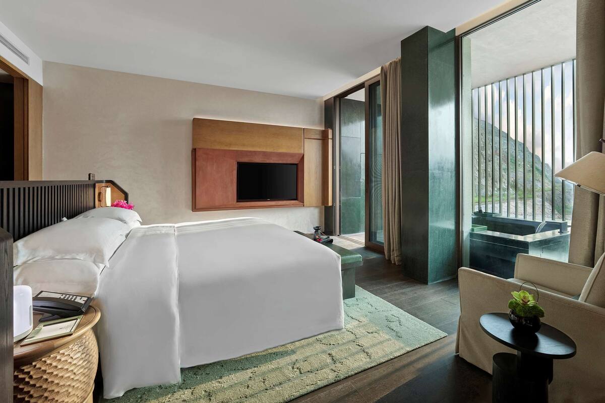 Een moderne hotelkamer met een groot bed, een flatscreen-tv aan de wand, een zithoek met een stoel en tafel, en uitzicht op een bergachtig landschap door grote ramen.