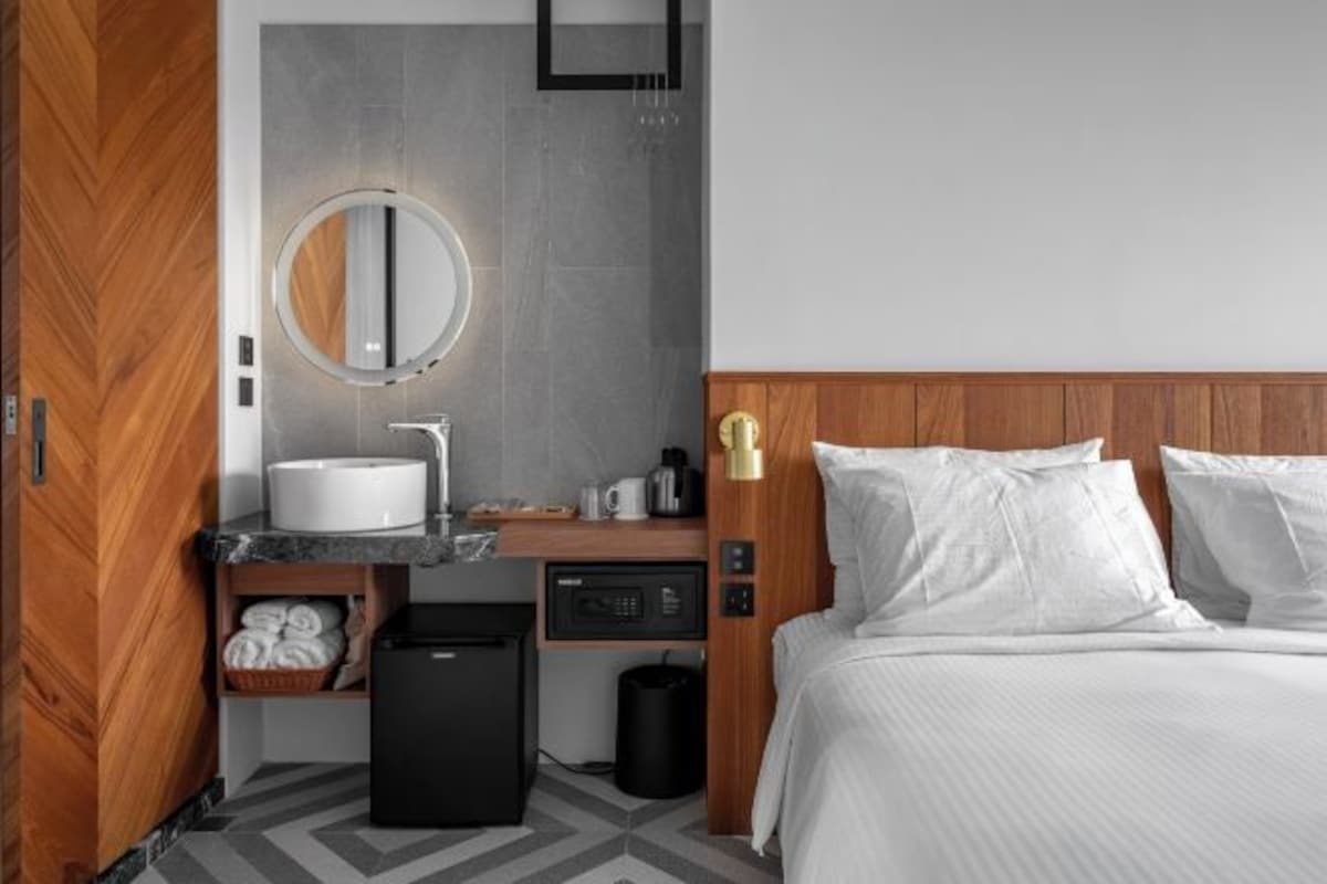 A modern hotel room with a wooden wardrobe, a round mirror, a sink, a compact refrigerator, a bedside table with a coffee maker, and a bed with white linens.