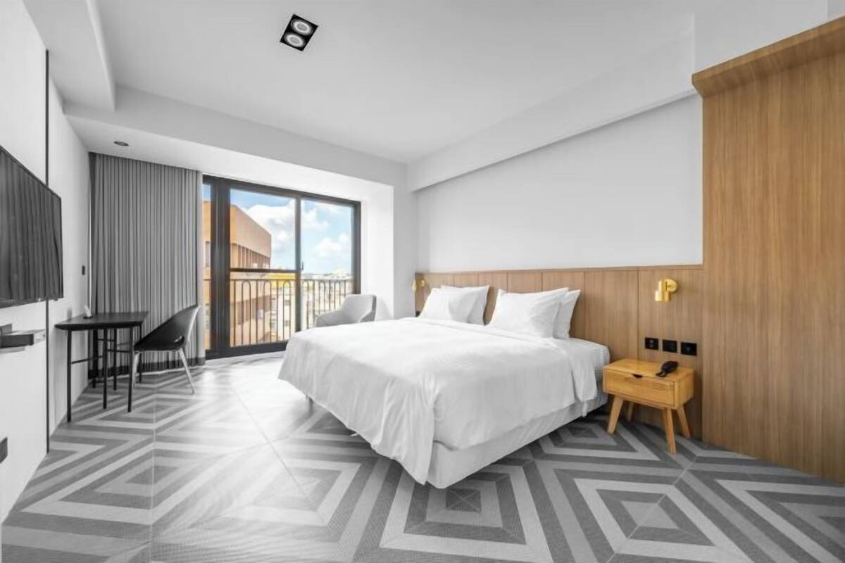 A modern hotel room with a large bed, a desk, and a chair, a wooden nightstand, and a view of the city through the window.