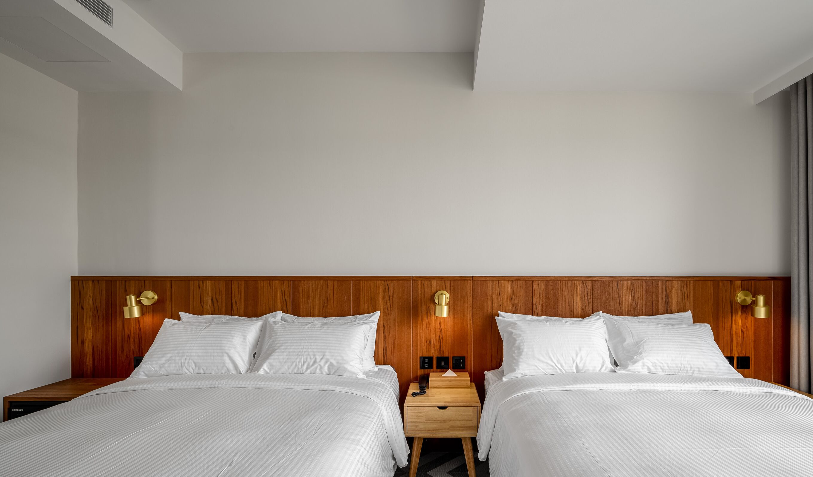 A hotel room with two beds, wooden headboards, bedside tables, and wall-mounted lamps.