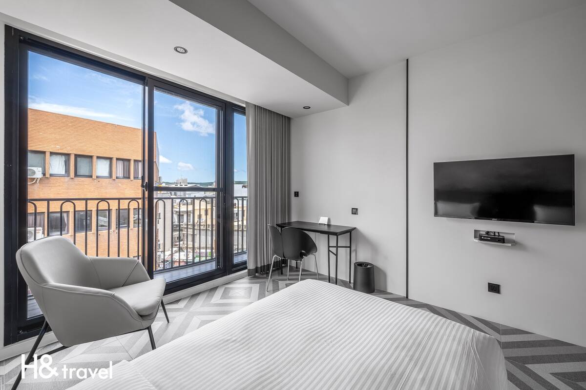 A modern hotel room with a large bed, a desk, a chair, a TV, and a balcony with a view of buildings.