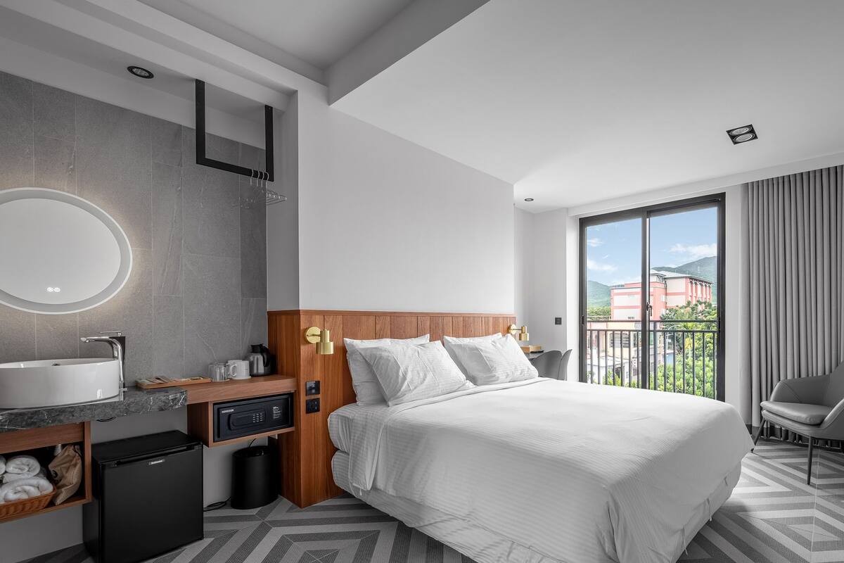 A modern hotel room with a large bed, a built-in desk, a round mirror, and a view of the outdoors.
