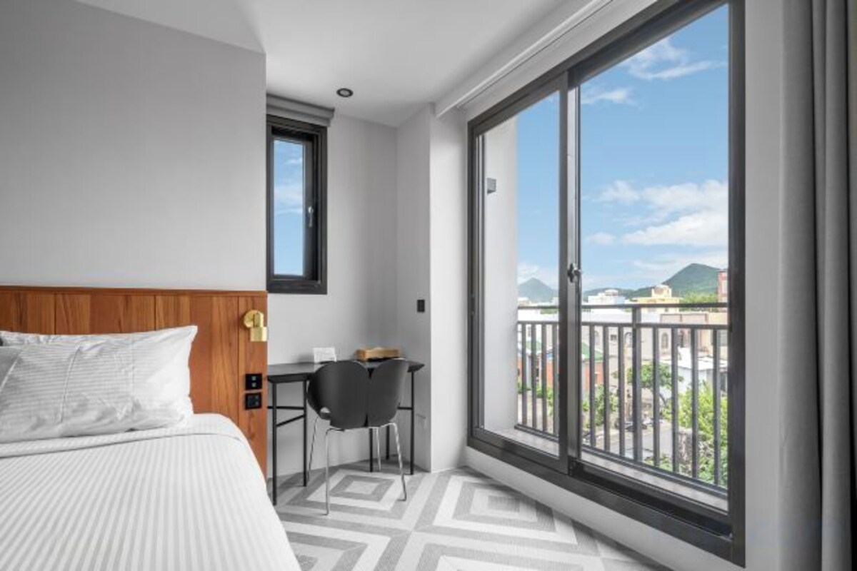 A hotel room with a bed, a desk, and a large window with a view of buildings and mountains.
