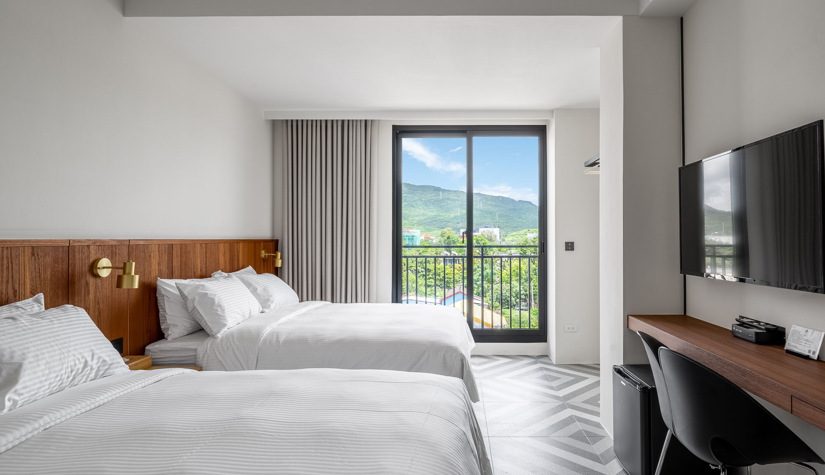 Habitación de hotel con dos camas, televisión, escritorio y balcón con vista a la ciudad.
