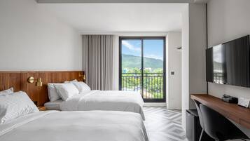 Habitación de hotel con dos camas, televisión, escritorio y balcón con vista a la ciudad.