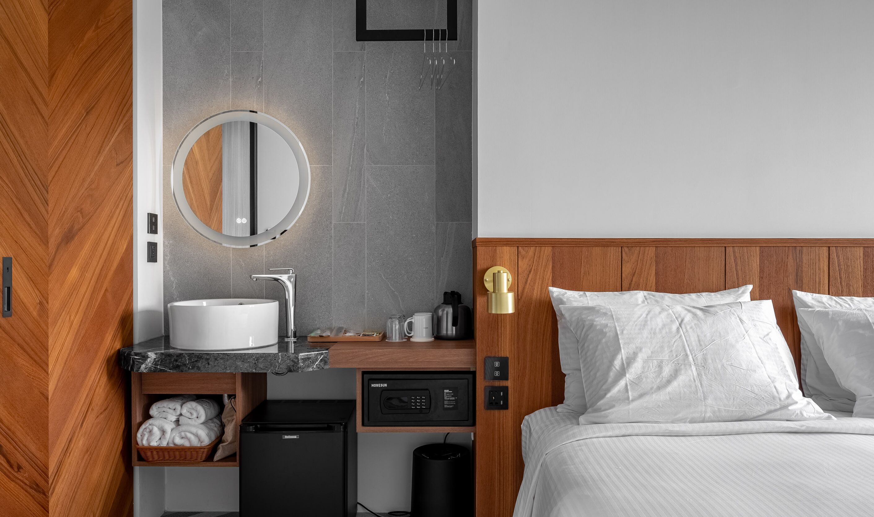 A modern hotel room with a wooden headboard, a round mirror above the sink, a built-in heater, a compact refrigerator, and a neatly made bed with white linens.