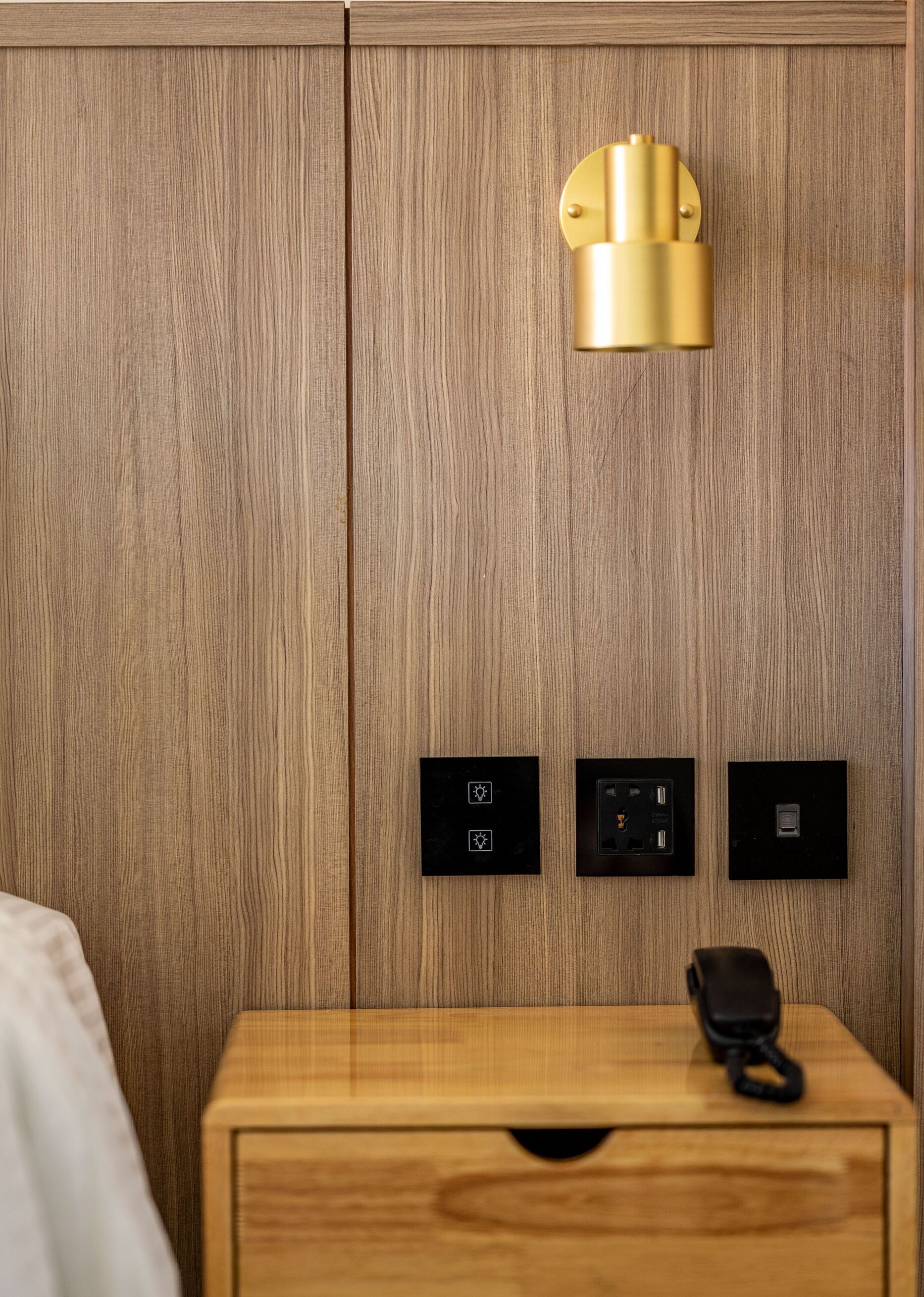 A wooden wall with a gold light fixture, three black switches, and a wooden nightstand with a black phone.