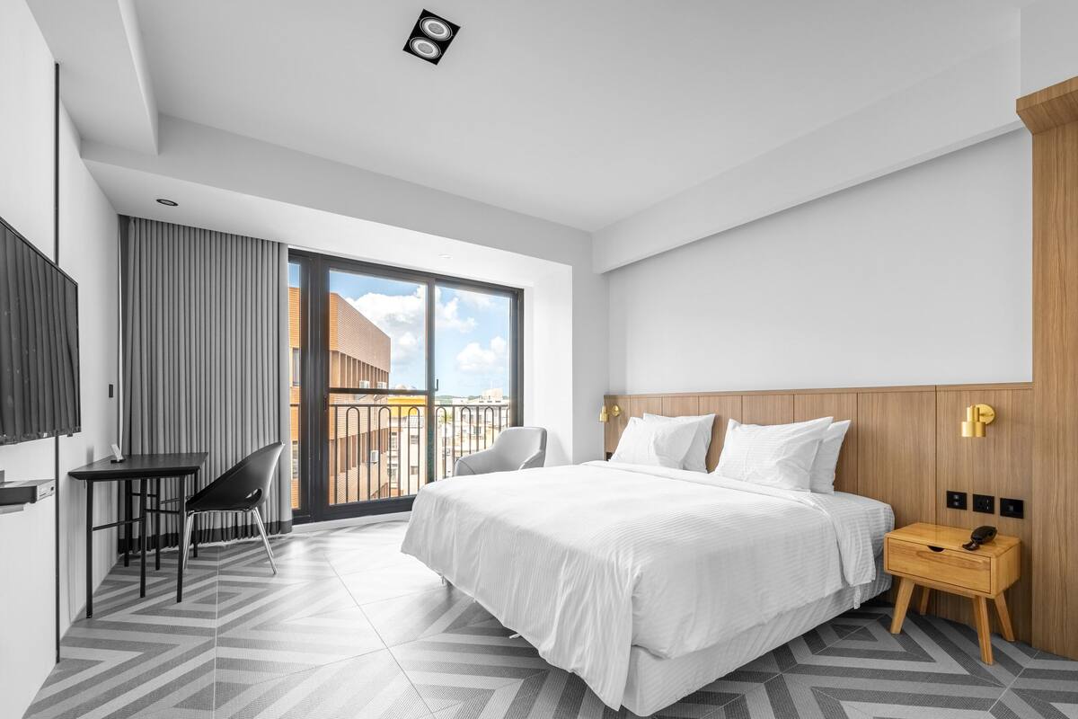 A modern hotel room with a large bed, a desk, a chair, and a view of the city through a balcony.