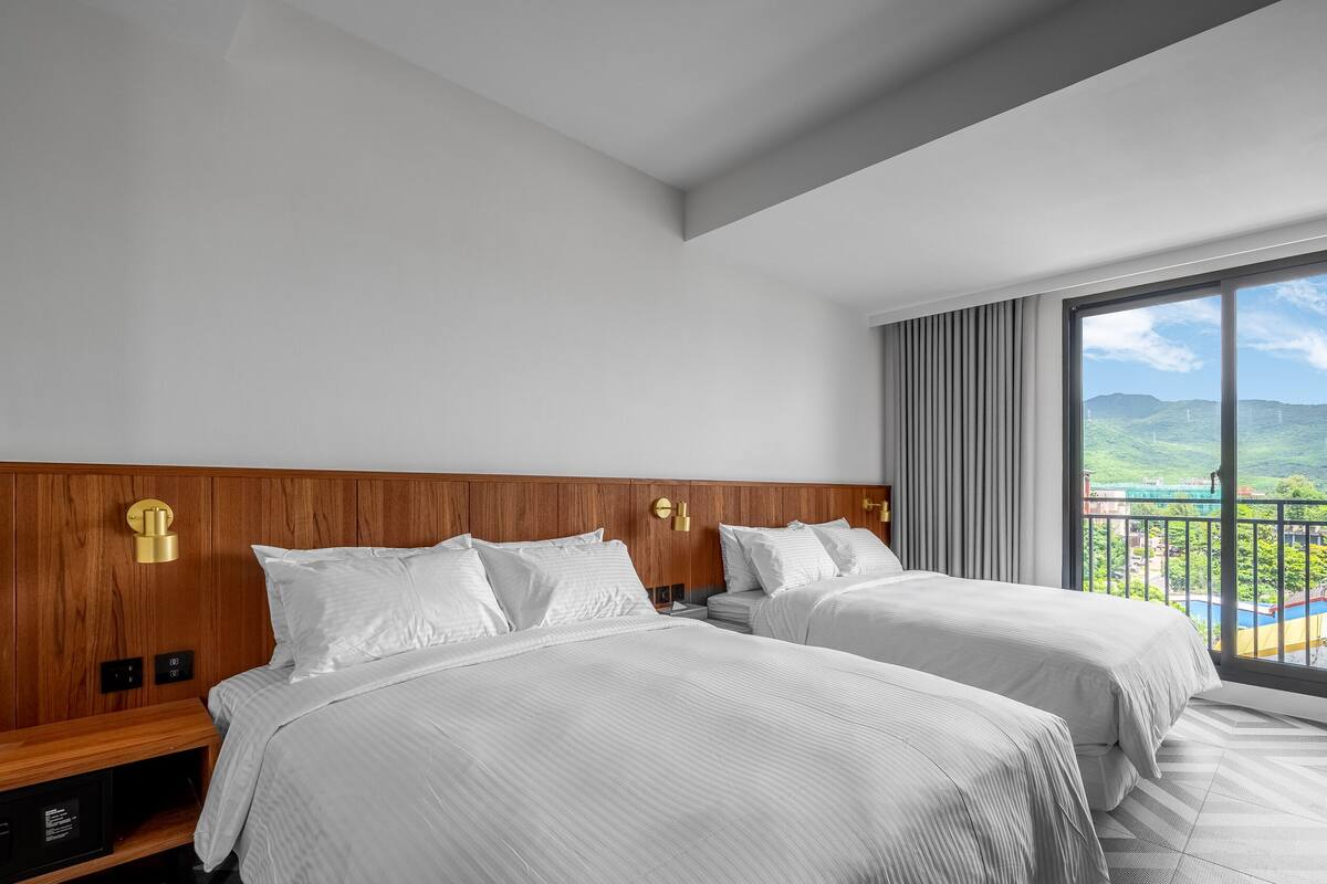 A hotel room with two beds, a wooden headboard, a large window with a view of hills, and a control panel on the left side.