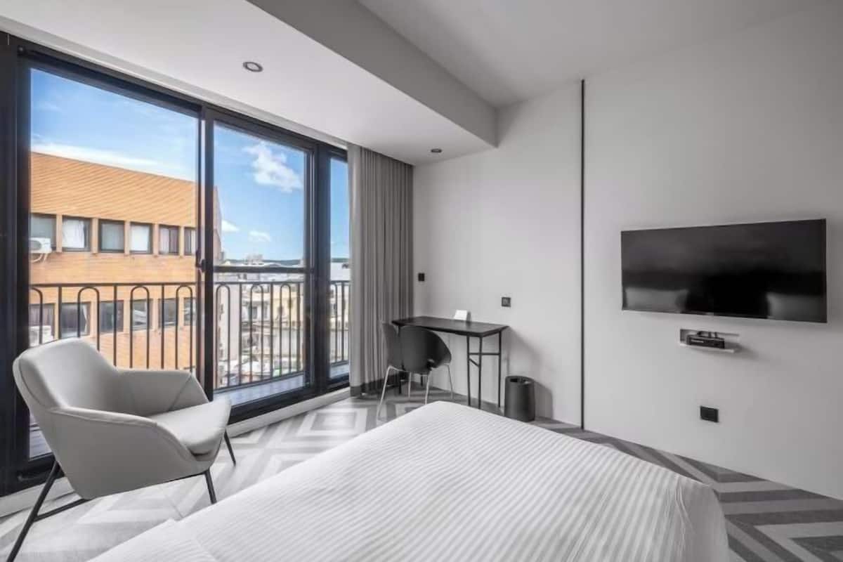 A hotel room with a bed, a desk, a chair, a TV, and a large window with a view of buildings.