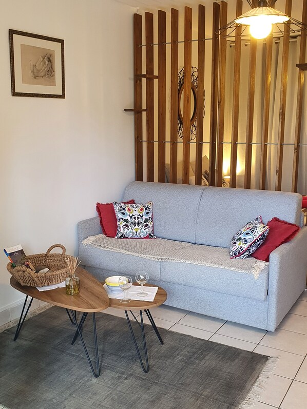 A living room with a grey sofa, two patterned pillows, a woven basket, a small table with a candle and glasses, and a wooden partition with vertical slats.