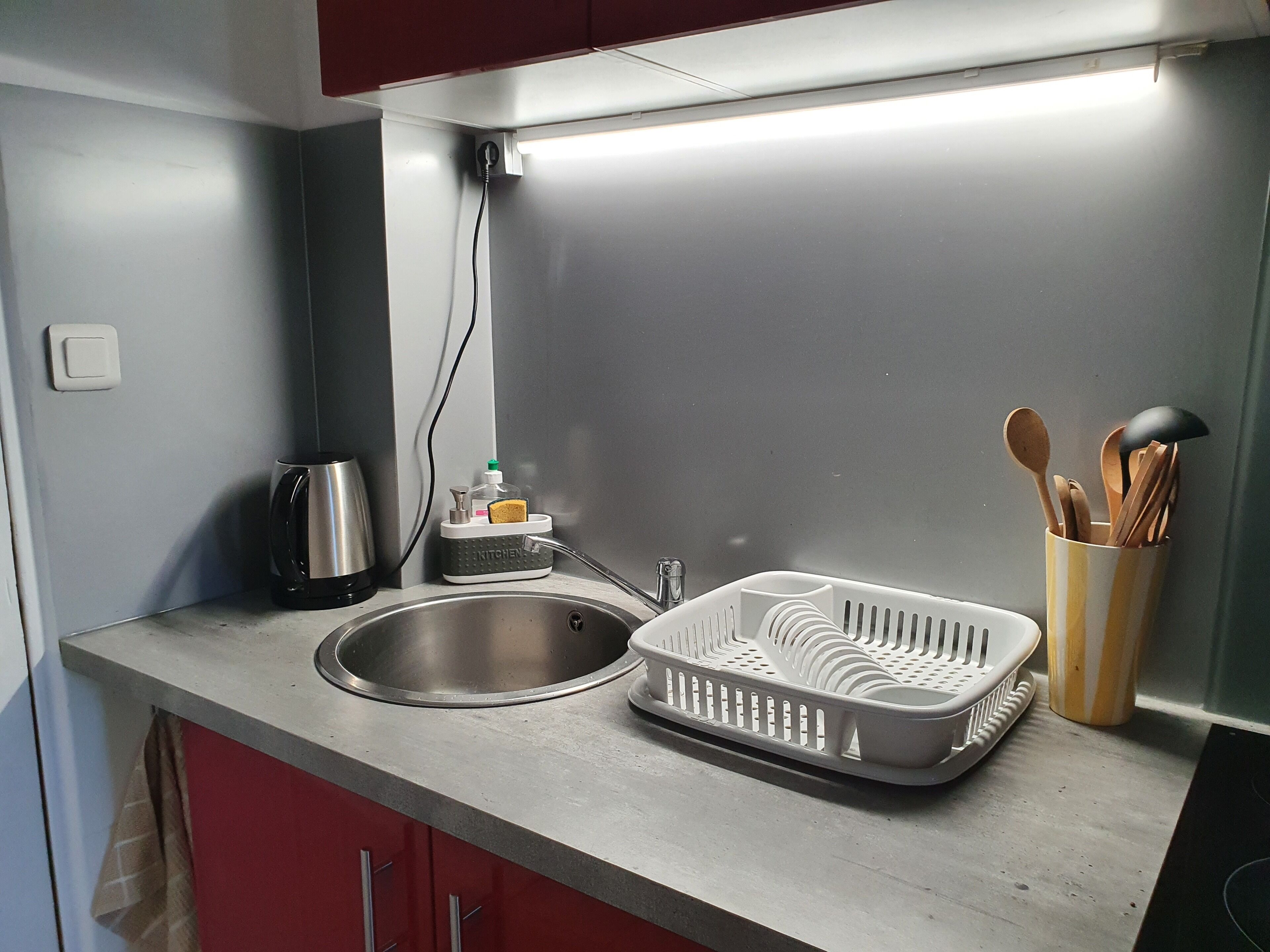 Et køkken med stålvask, elkedel, opvaskestativ og bestikholder.