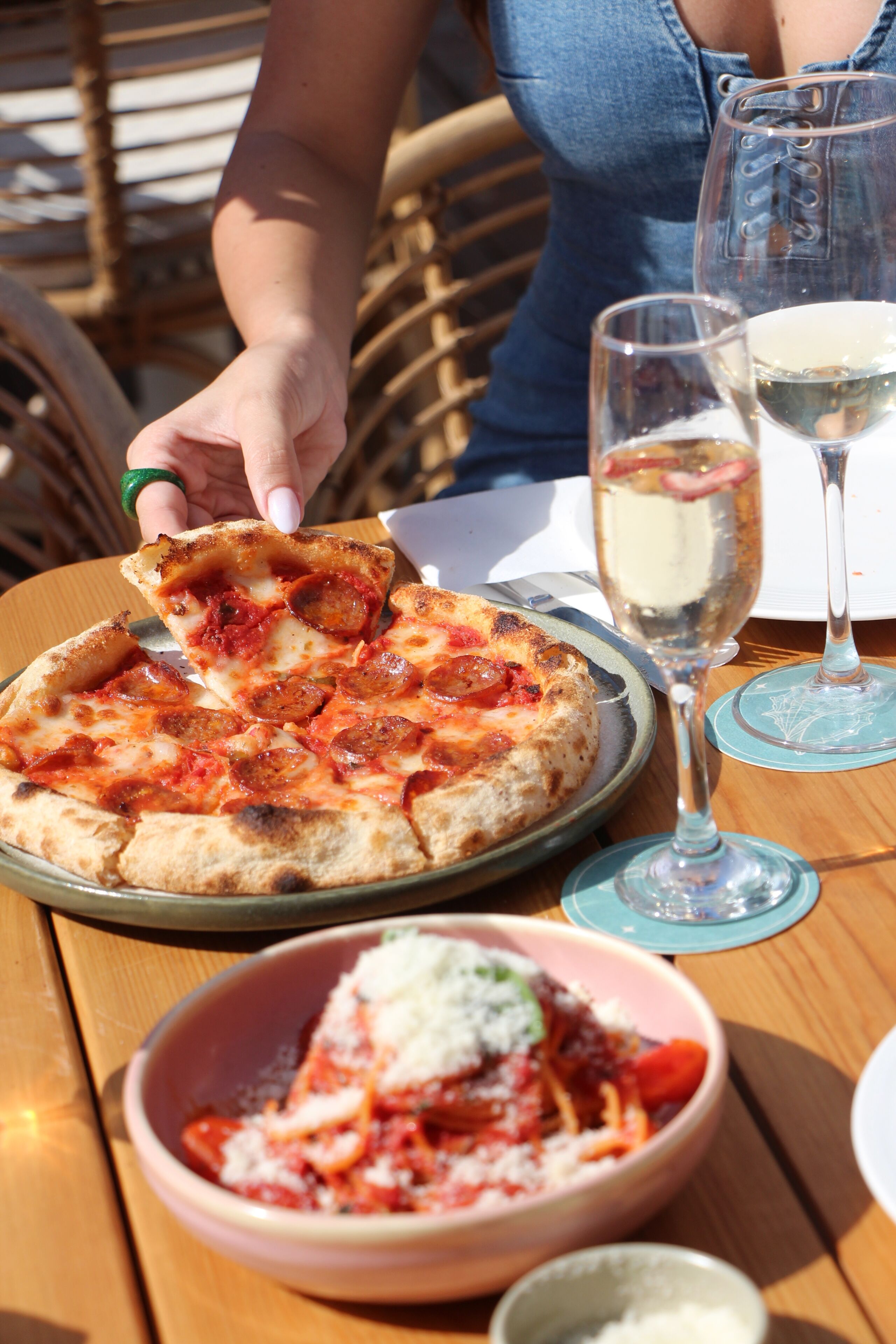 Una pizza en un plato con una porción que se levanta, una copa de vino blanco y un plato de pasta con queso sobre una mesa.