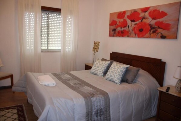 A bedroom with a bed, a nightstand, a lamp, a painting, and a window with curtains.