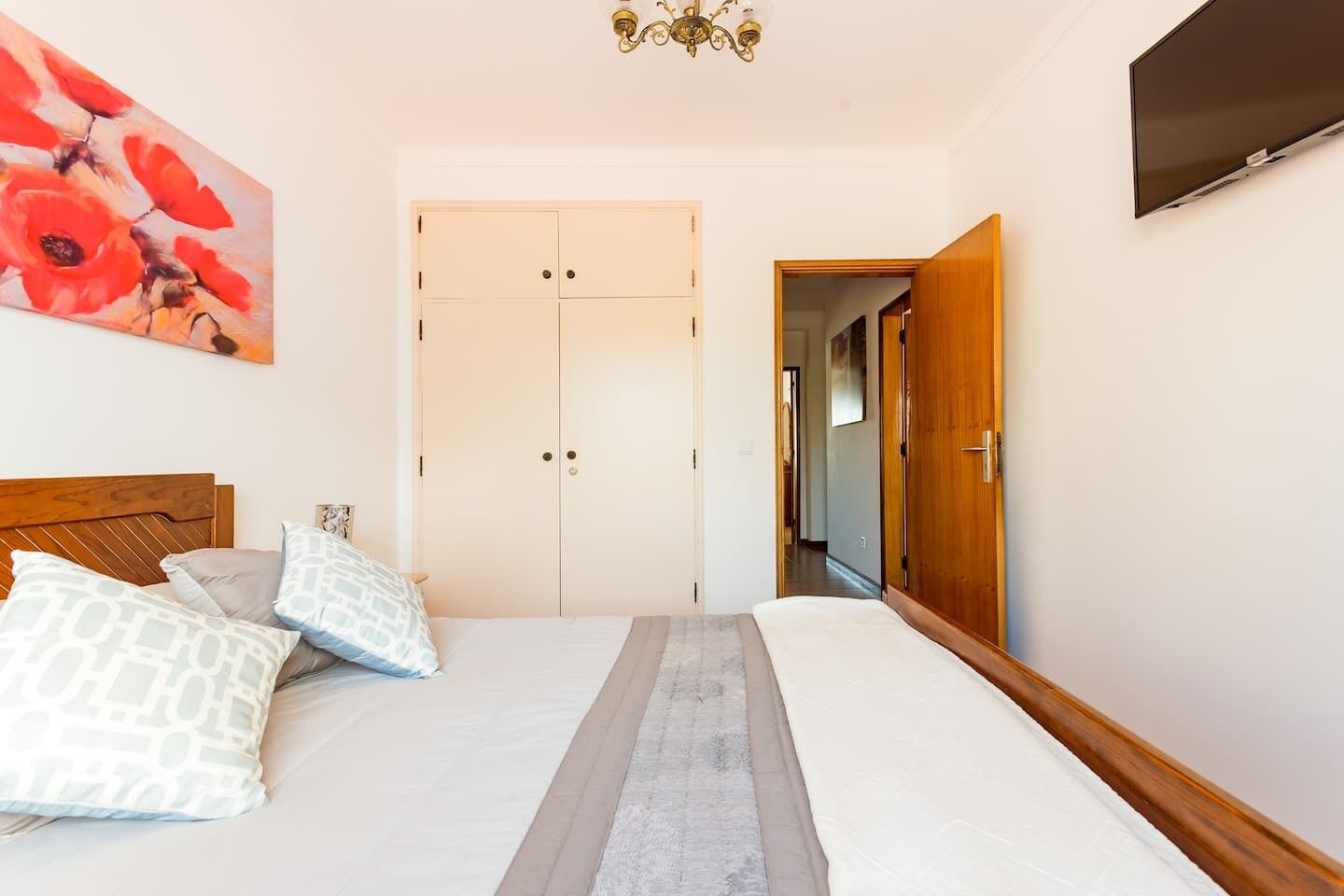 A bedroom with a bed, a wooden headboard, a painting, a wardrobe, and a TV on the wall.