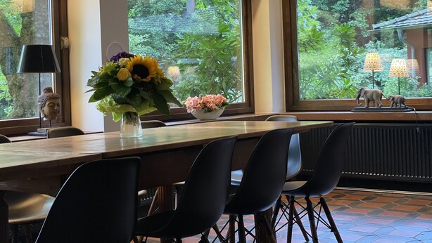 A dining area with a long table, black chairs, a Buddha head sculpture, a vase with flowers, and a view of greenery and a building through large windows. - House (Lohmar)