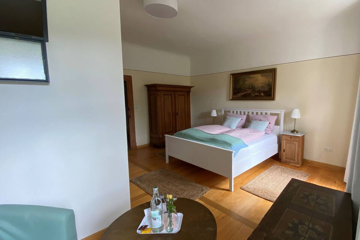 Un dormitorio con una cama individual, una mesita de noche, un armario de madera, un cuadro en la pared y una pequeña mesa con una botella y vasos.