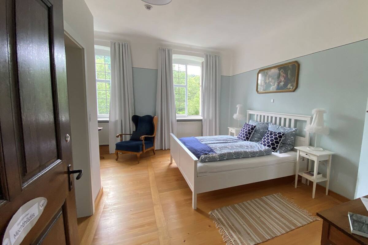 Un dormitorio con una cama, una silla, una mesita de noche, un reloj y una ventana.