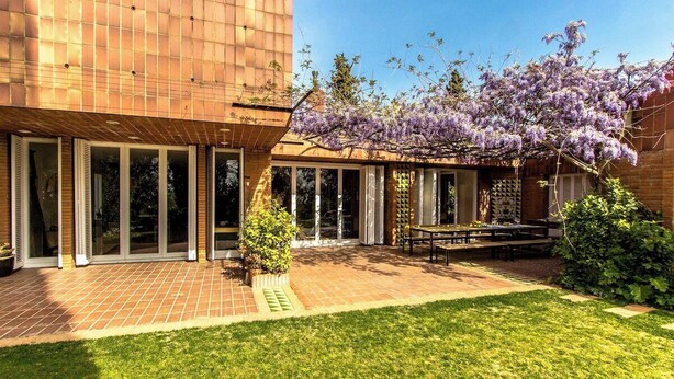 Een modern gebouw met een met baksteen bestraat terras, een overdekte zitplaats en een boom met paarse bloemen.