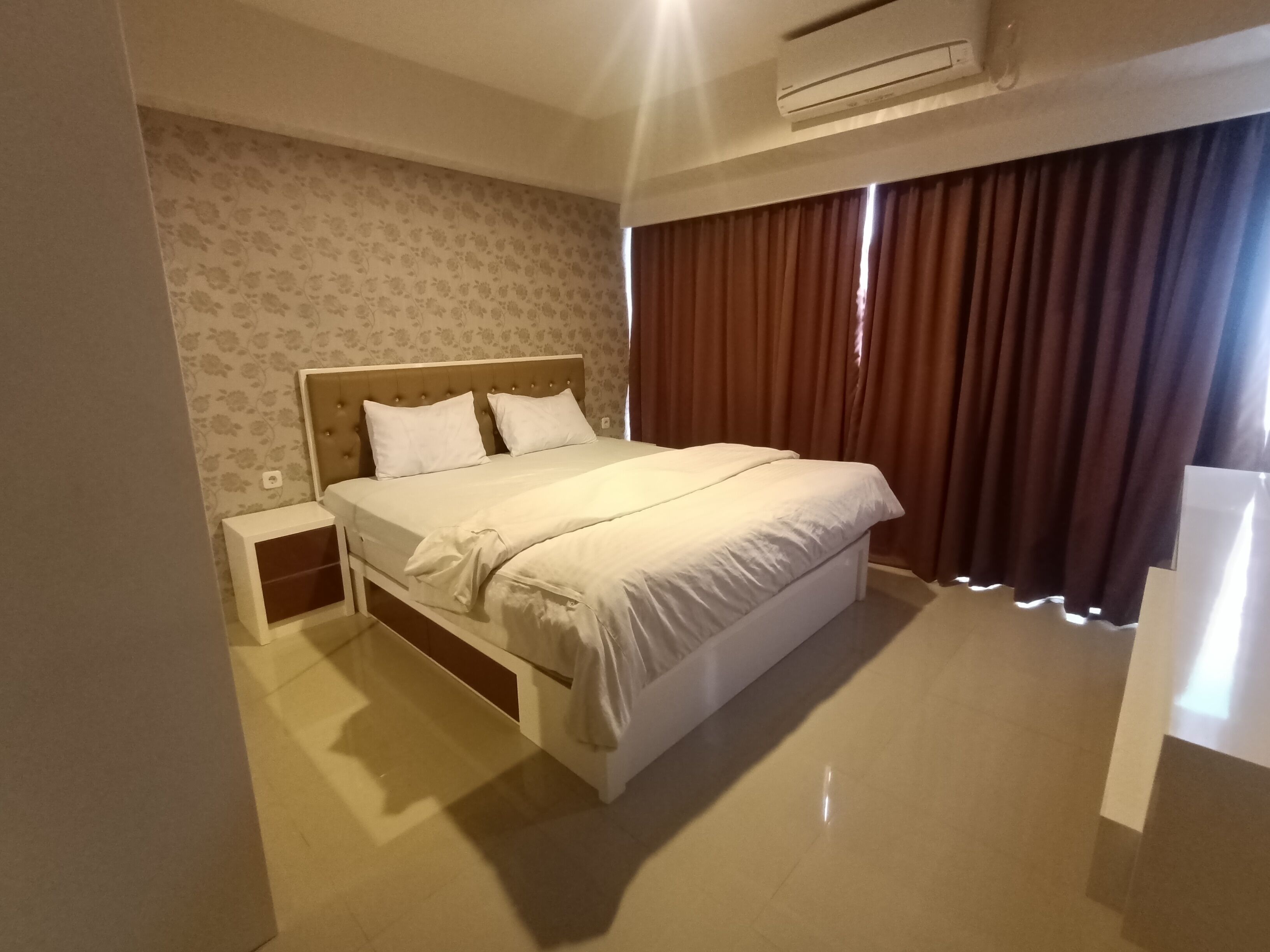 A hotel room with a bed, a nightstand, a wall-mounted air conditioner, and a window with curtains.