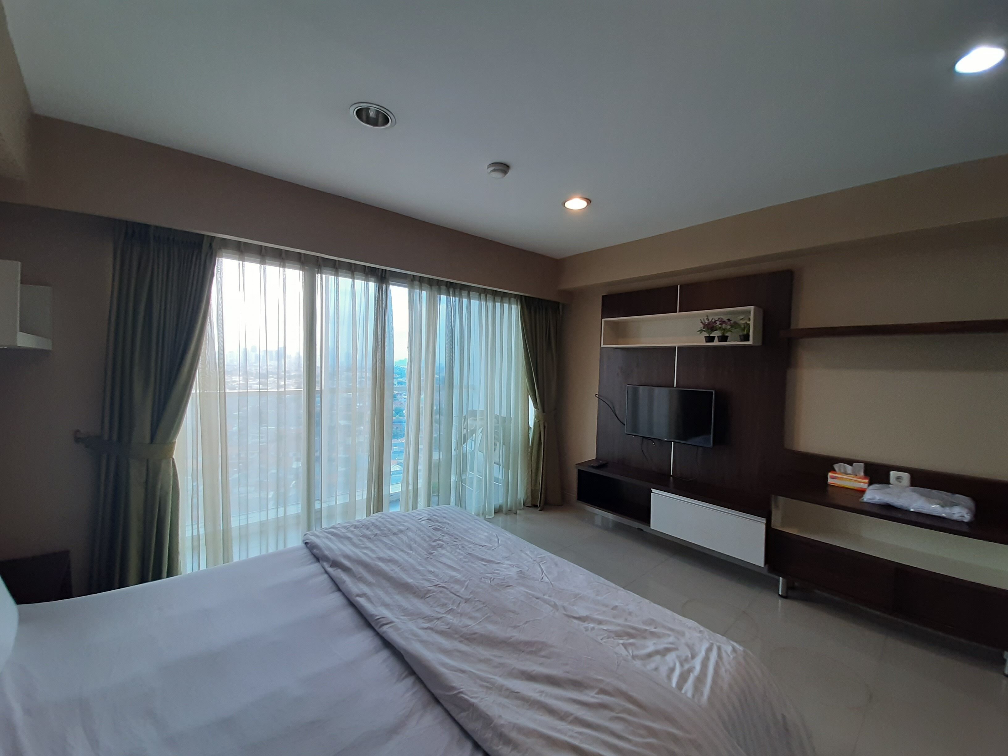 Habitación de hotel con una cama grande, un televisor de pantalla plana en la pared, un estante con una planta y una ventana con cortinas.