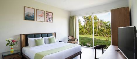 A modern hotel room with a large bed, a desk with a flat-screen TV, a ceiling fan, and a view of a tropical garden through large windows.