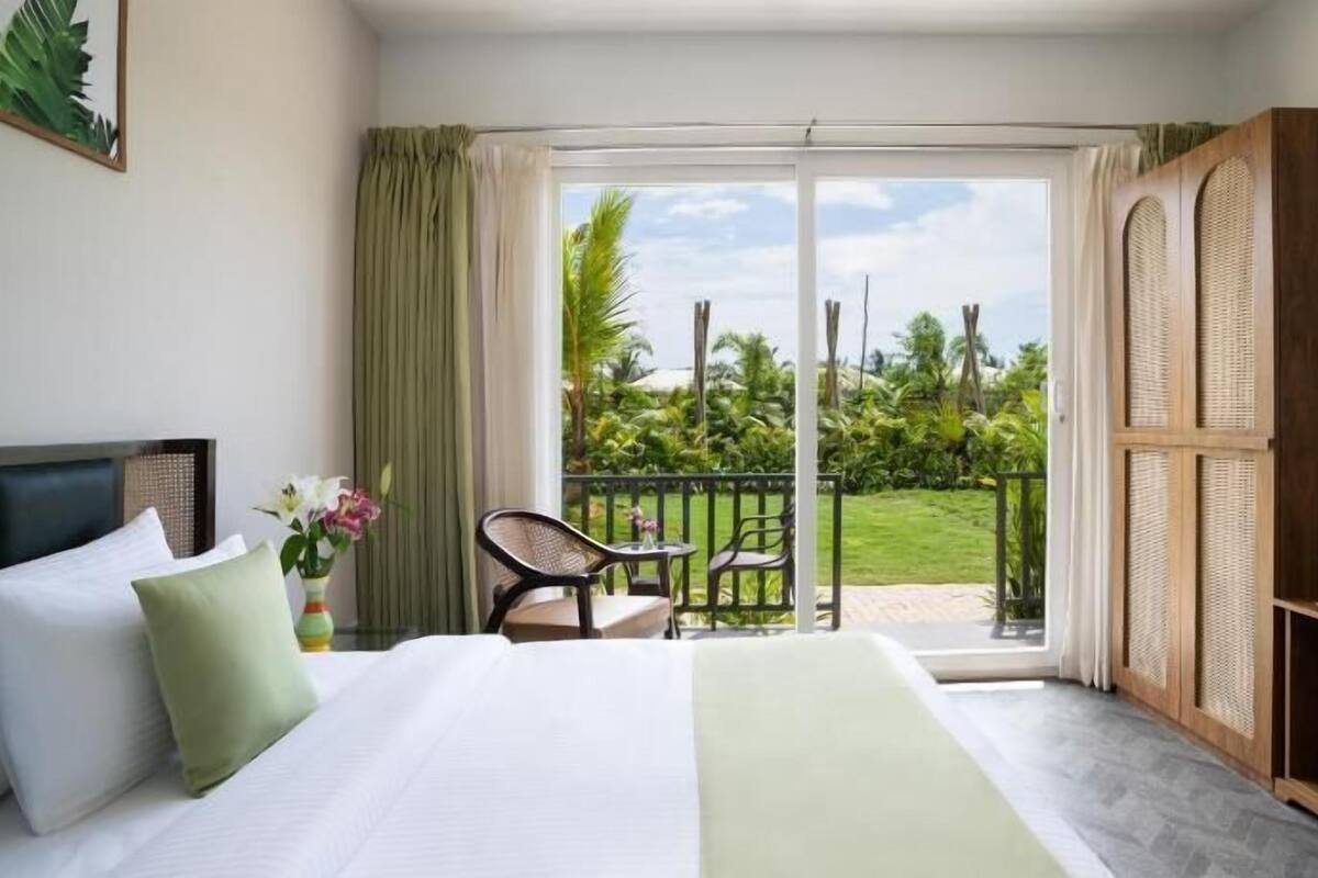Habitación de hotel con cama, una silla, una mesita y un balcón con vistas a la vegetación.