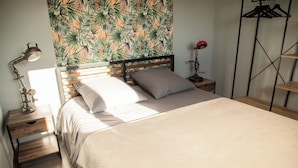 A neatly made bed with a headboard, a nightstand with a lamp, and a wall with a tropical leaf pattern. - LA TRANQUILITE NORMANDE Country cottage surrounded by animals! (Livarot-Pays-d'Auge)