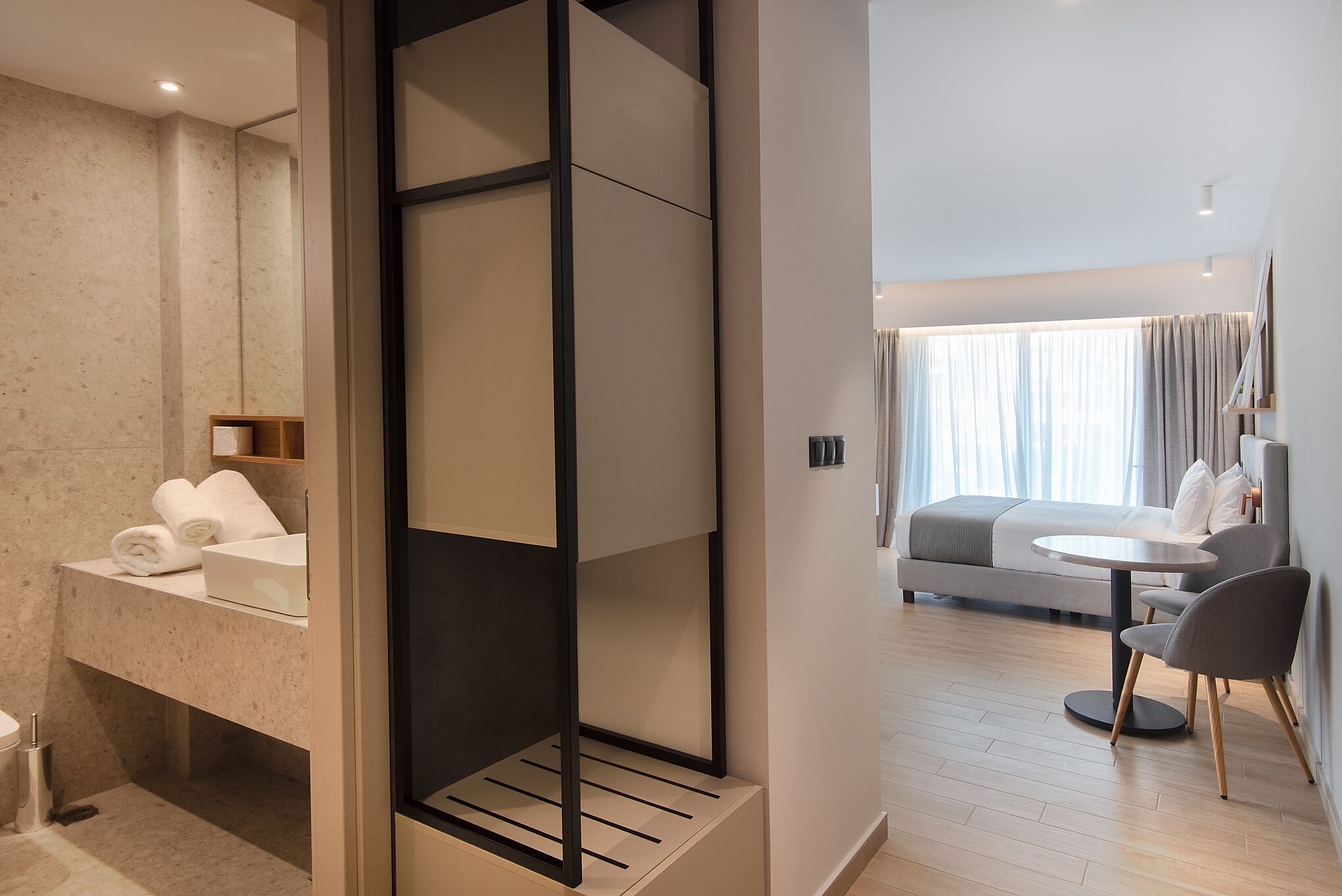 A modern hotel room with a bathroom, a bed, a dining area, and a large window with curtains.