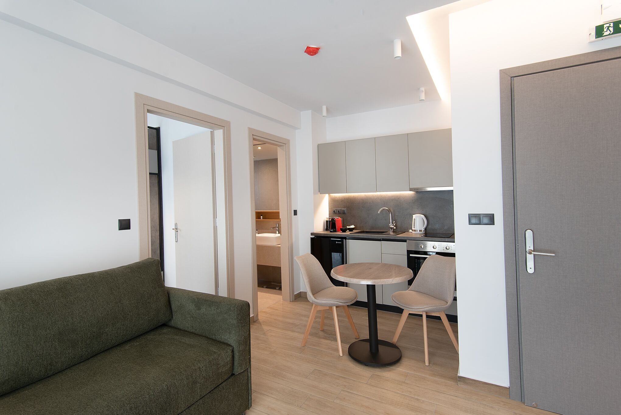 Una habitación de hotel moderna con un sofá verde, una mesa de comedor redonda con dos sillas, una kitchenette y una puerta gris.