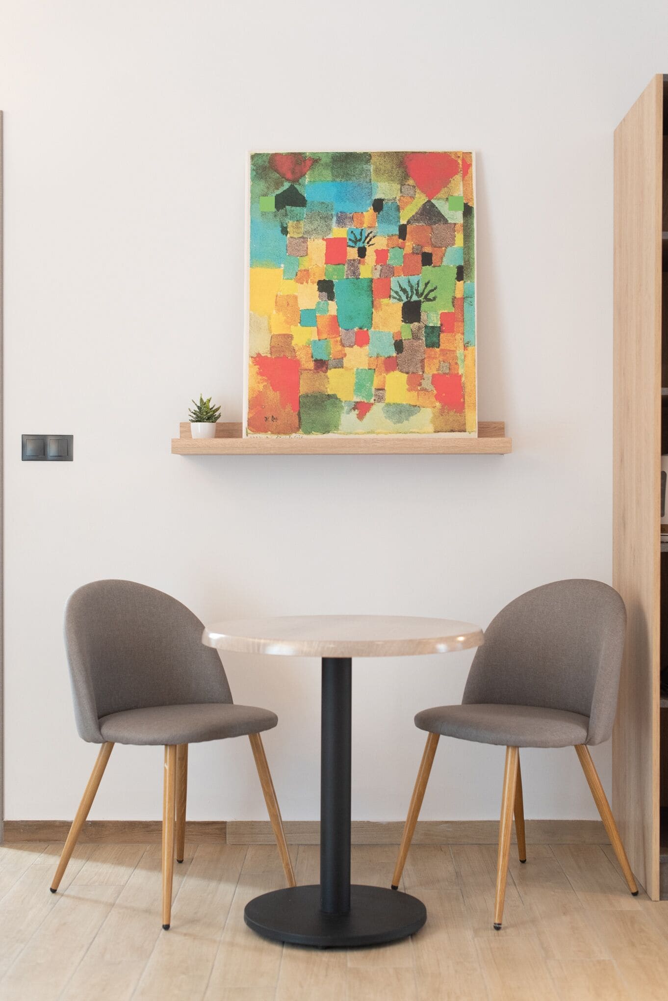 A modern room with a round table, two grey chairs, a colorful abstract painting on the wall, and a small plant on a shelf.