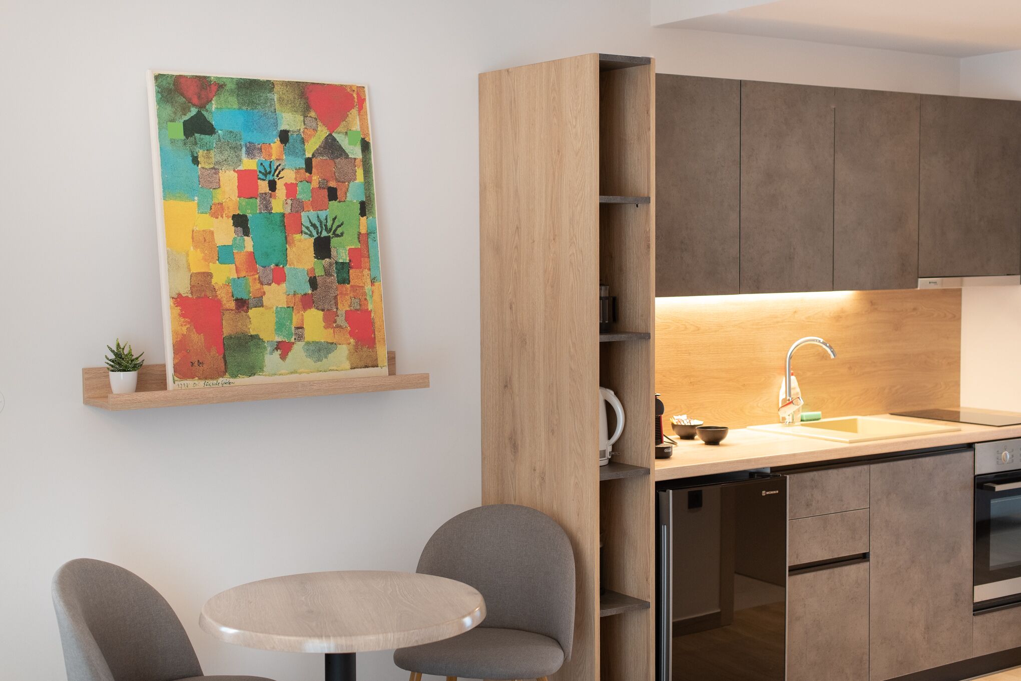 Una cocina moderna con armarios de madera, lavavajillas empotrado y un lavamanos. En la pared, arriba de una pequeña planta en maceta, hay un cuadro abstracto y colorido.