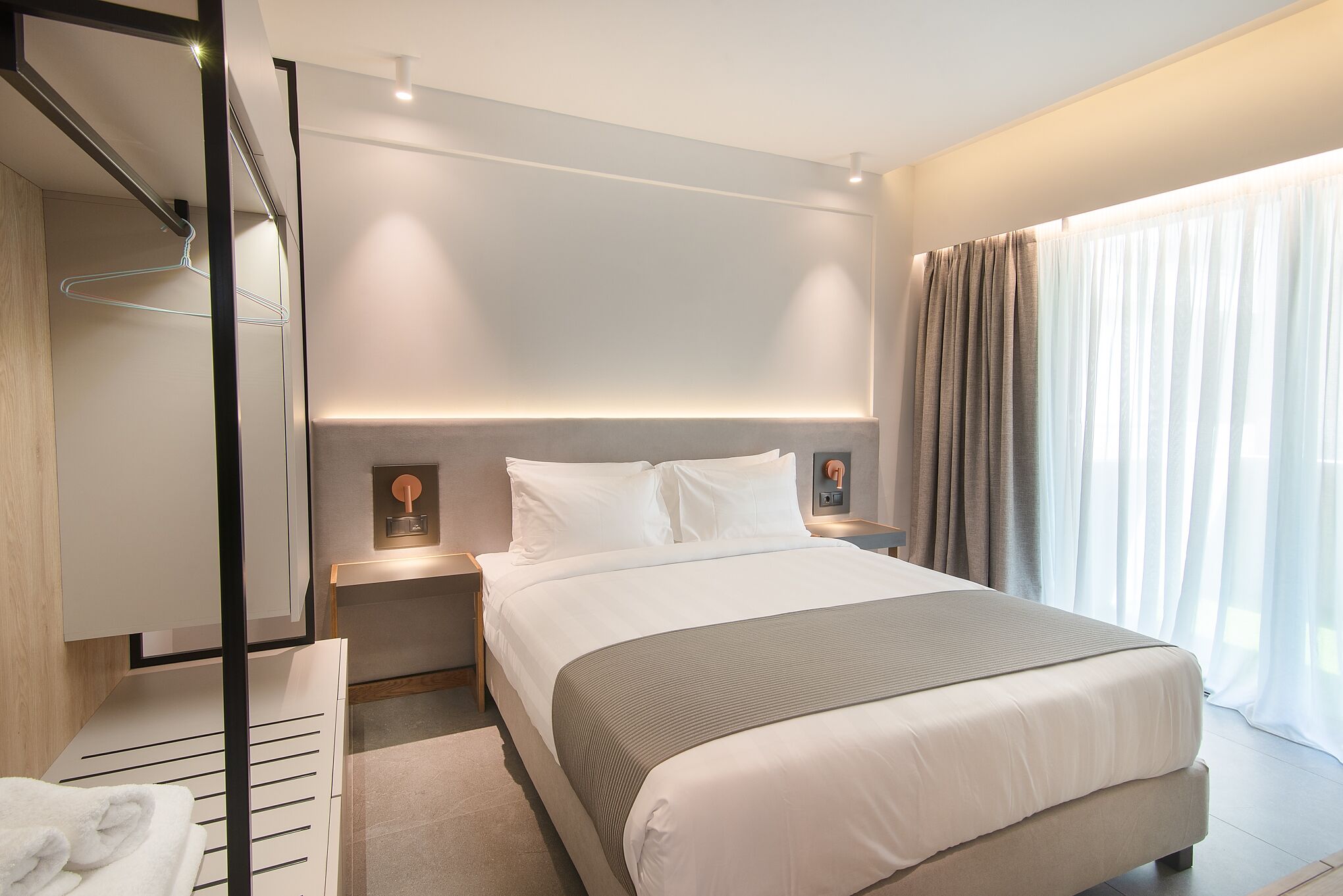 A modern hotel room with a large bed, bedside tables, a hanging light, and a view of the outdoors through sheer curtains.