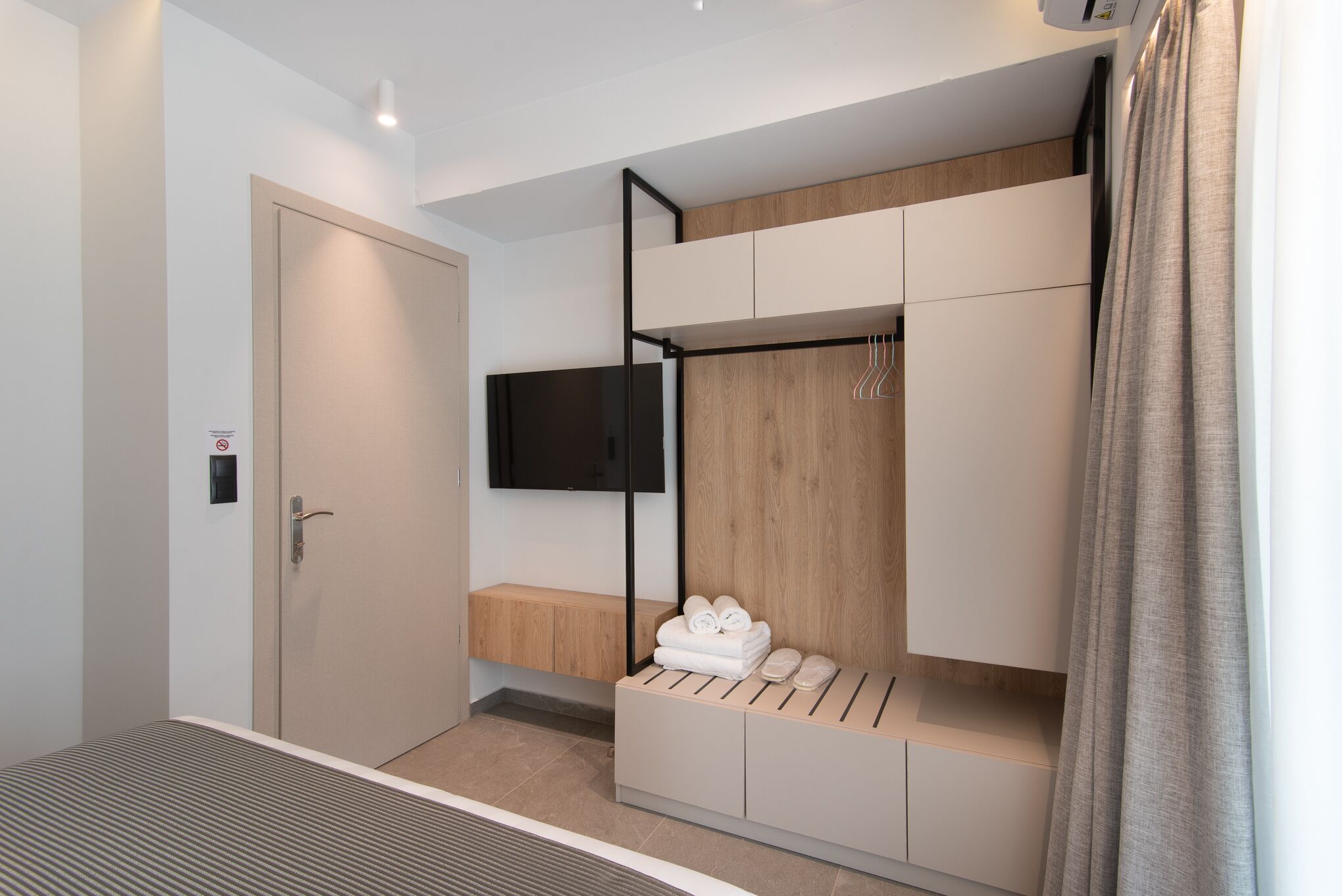 A modern hotel room with a bed, a TV mounted on the wall, a wooden bench, and a storage unit with towels.