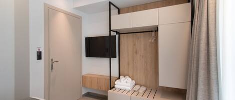 A modern hotel room with a bed, a TV mounted on the wall, a wooden bench, and a storage unit with towels.