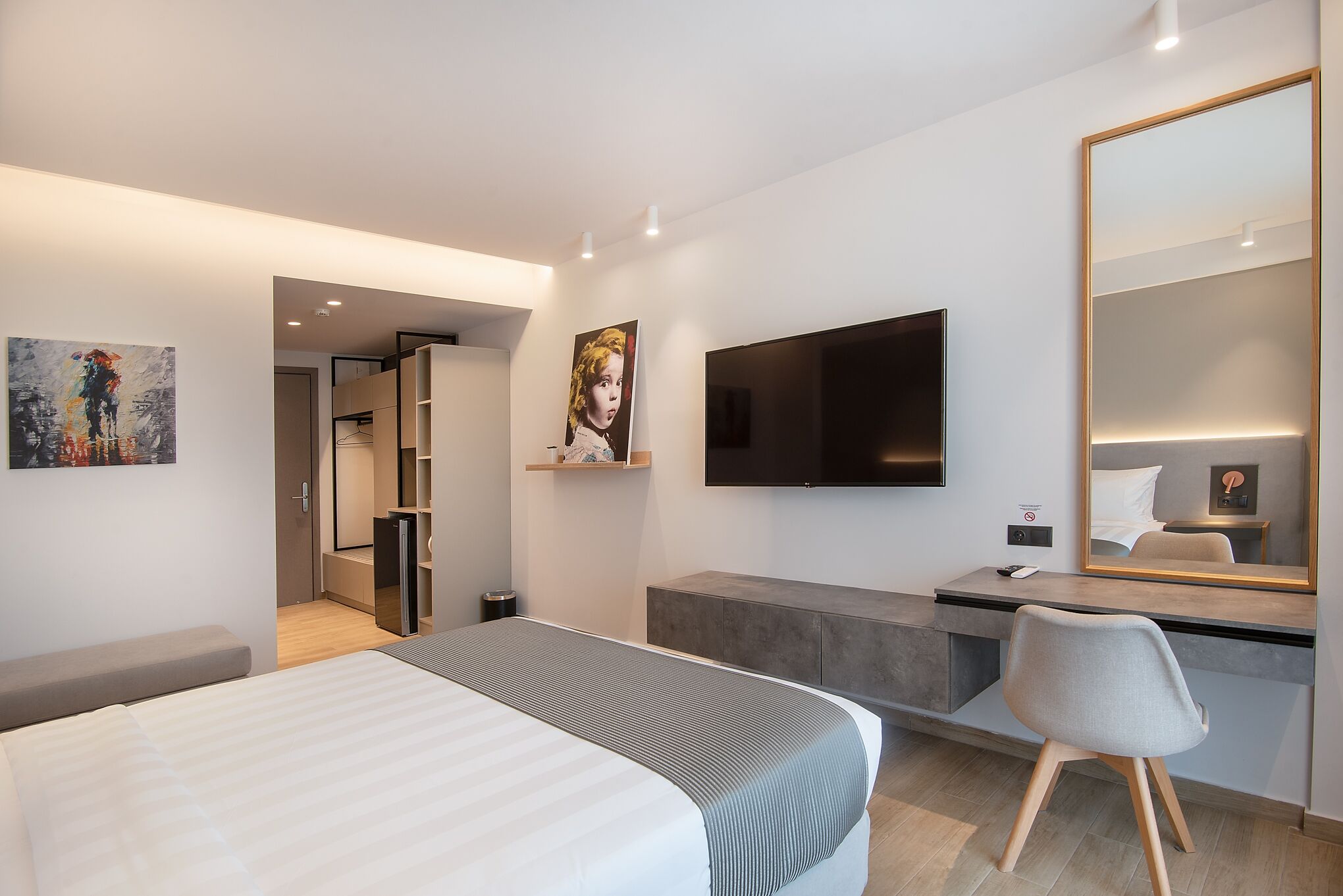Ett modernt hotellrum med en säng, ett skrivbord, en stol, en TV och konstverk på väggarna.