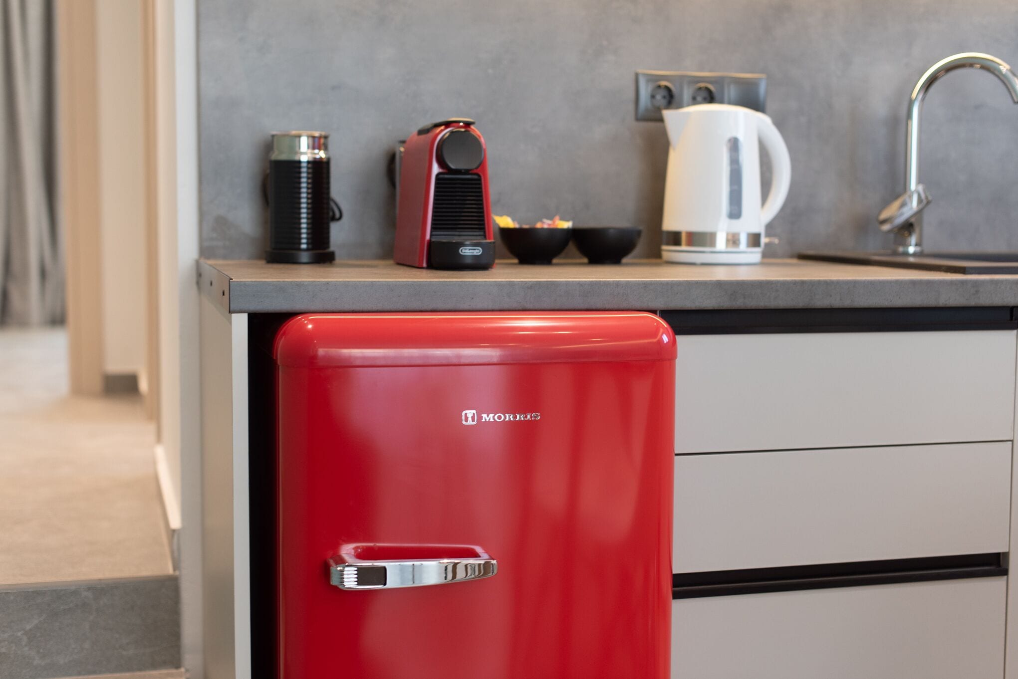 Una cucina compatta con un elettrodomestico rosso, una macchina da caffè e un bollitore sul piano di lavoro.