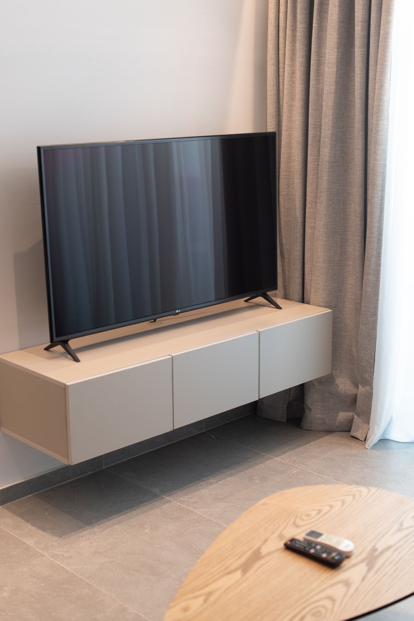 Un televisor de pantalla plana sobre un mueble blanco, un control remoto sobre una mesa de madera y una cortina al fondo.