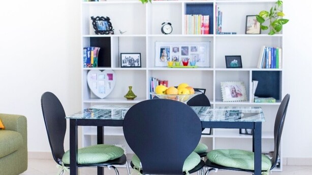 A dining area with a glass-top table, black chairs with green cushions, and a white bookshelf in the background. - apartment with terrace (Sant'Antioco)