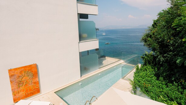A modern hotel with a glass infinity pool overlooking the sea, a clear sky, and a sign for the Pay Hotel Beach & Restaurant. -  Ray Hotel Corfu (Corfu)