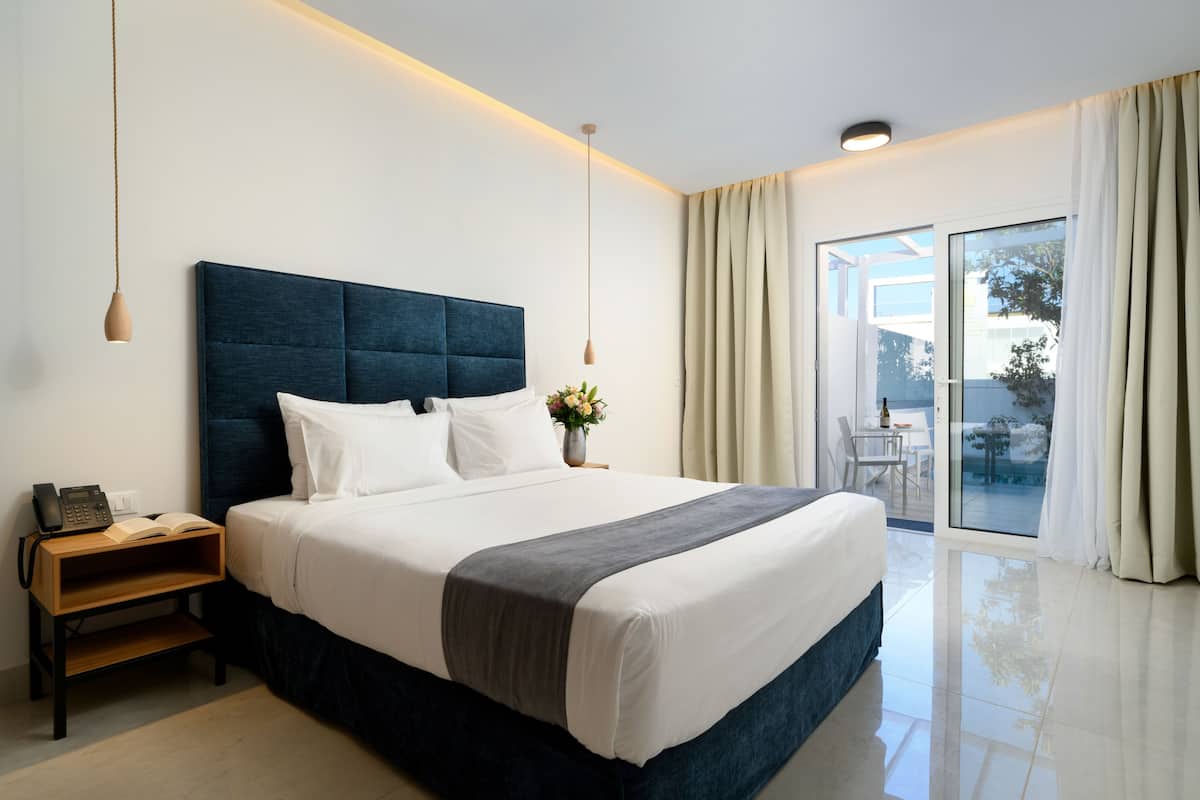 Una habitación de hotel moderna con una cama grande, mesitas de noche, una pequeña mesa con un florero y una puerta corredera de vidrio al balcón.