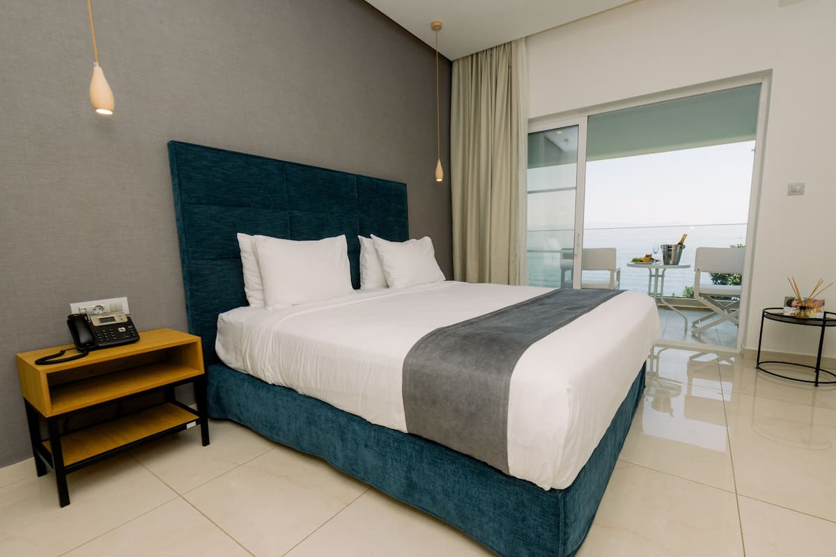 Una habitación de hotel moderna con una cama grande, una mesita de noche con teléfono, una pequeña mesa con una bebida y vistas al mar.