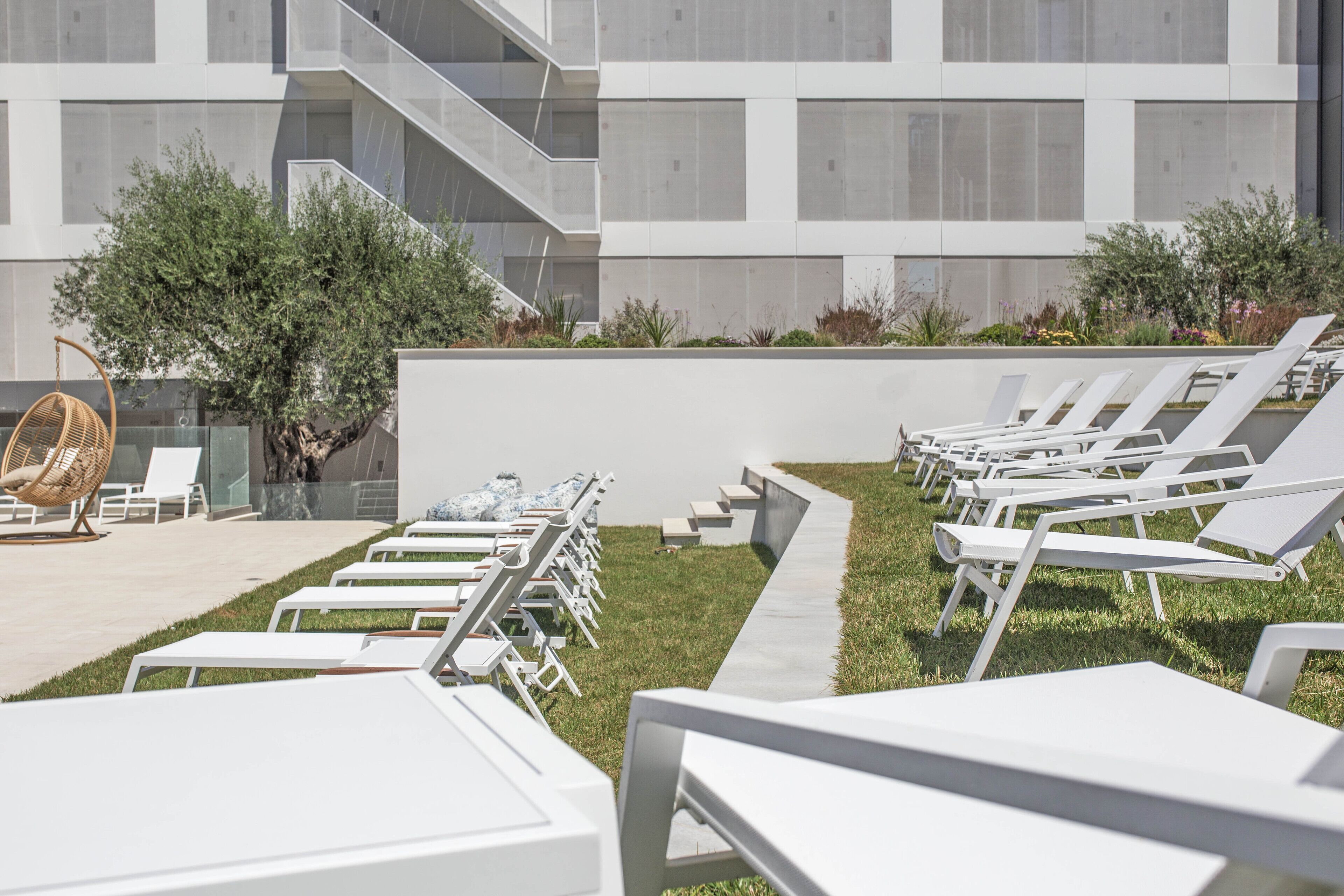 A modern outdoor lounge area with white sun loungers and a wicker chair, a staircase, and a building with a geometric facade.