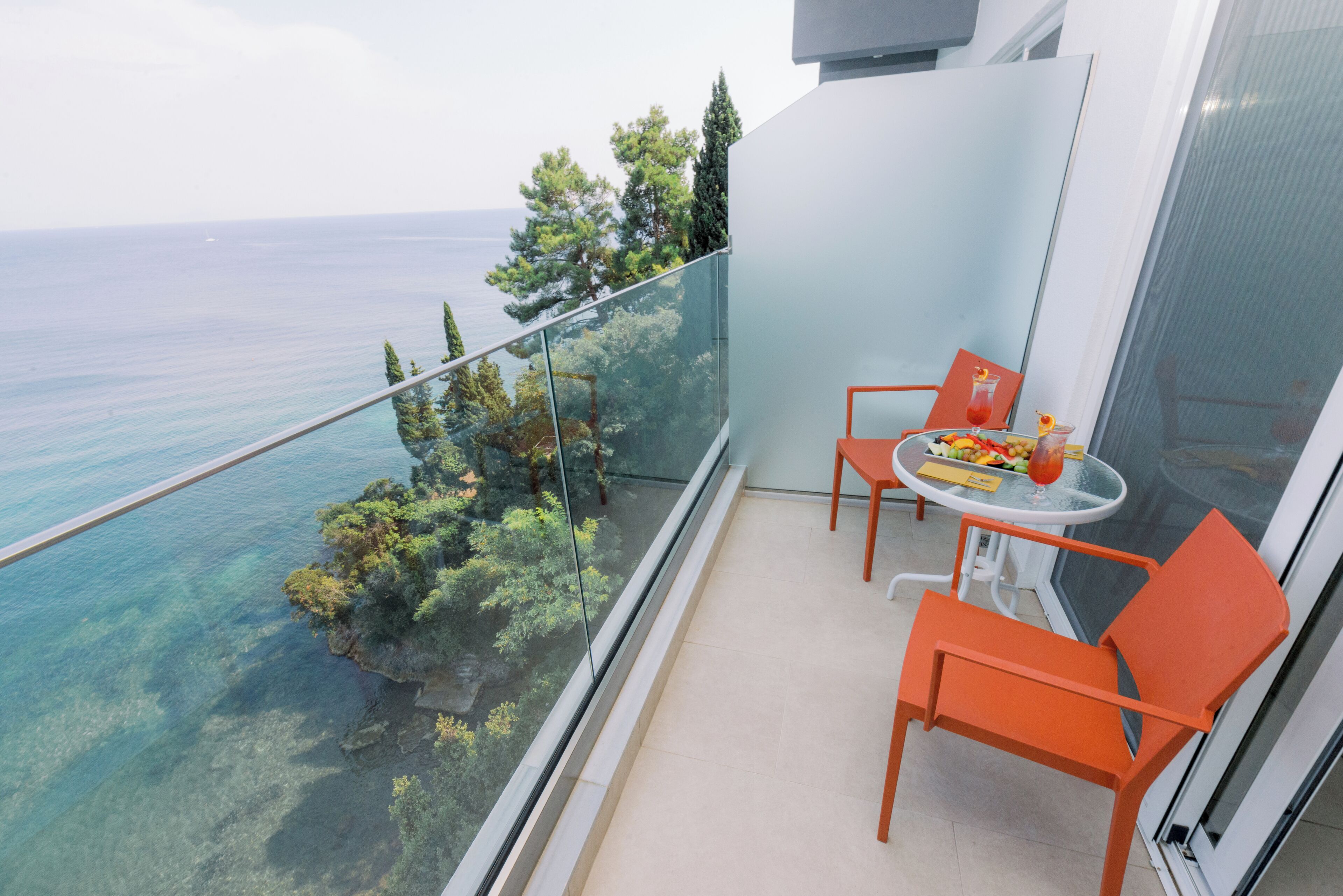 A balcony with a glass railing overlooking a coastal area with trees and the sea. There is a table with orange chairs and a fruit bowl.